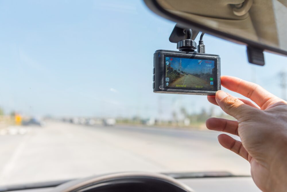 Dash cam mounted on windshield of vehicle. Hand adjusting position. Highway in the background.