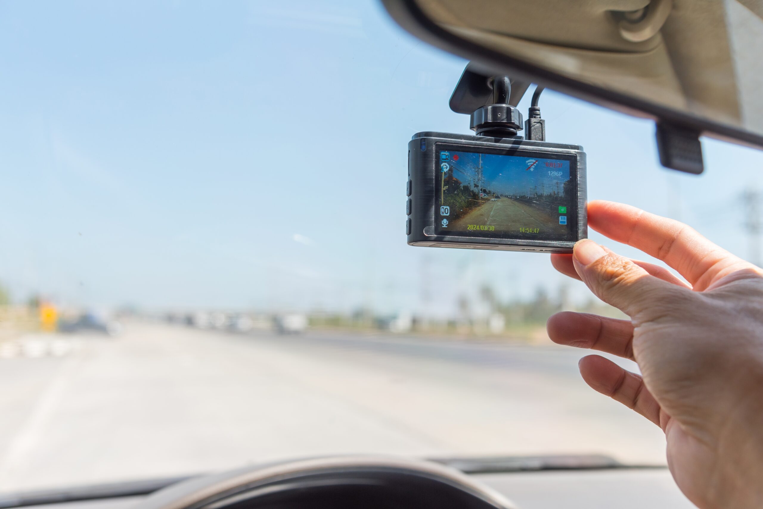 Dash cam mounted on windshield of vehicle. Hand adjusting position. Highway in the background.