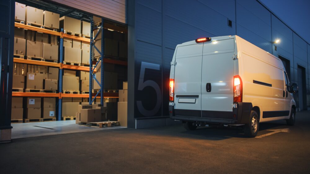 White delivery van parked outside a distribution center open door. Boxes seen inside warehouse.